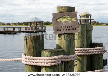 A wooden sign posted on a pier or wharf warning people to please no fishing in the area. The wooden signage has a graphic of a fish and black text. There's water and a boat in the background.