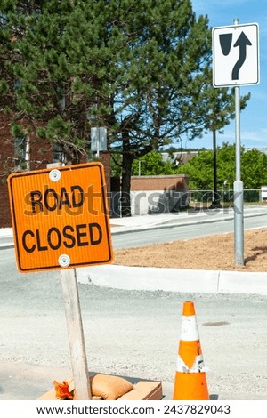 An orange colored road sign with a road closed in black lettering. The sign is in the foreground with a road construction site in the background and a large brick building with tall green trees.