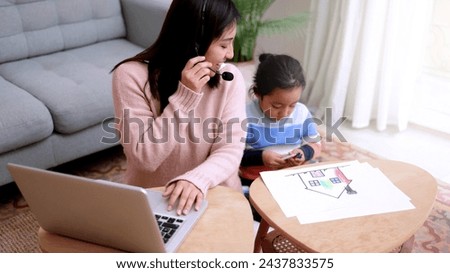 Asian mother work on computer at home with child son. Busy mom giving call center support Royalty-Free Stock Photo #2437833575