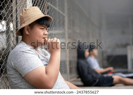 Asian teenboy in white t-shirt sits with picking teeth against a metal fence panel in a juvenile detention facility, awaiting further release, freedom and detention of people concept. Royalty-Free Stock Photo #2437855627