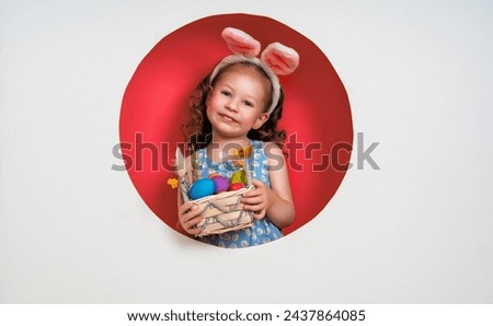 Cute little child wearing bunny ears on Easter day. Girl with painted eggs on white and pink background. Royalty-Free Stock Photo #2437864085