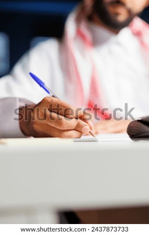 A focused businessman of Middle Eastern ethnicity efficiently works at his home office desk, taking research notes using a blue pen. Selective focus an Arab person writing down on his notebook. Royalty-Free Stock Photo #2437873733