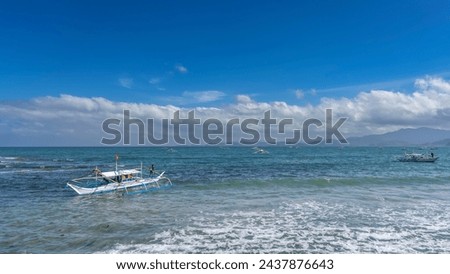 A traditional Filipino   boat bangka with a double-outrigger dugout bobs on the waves of the turquoise ocean. On the horizon, against the blue sky and clouds, a mountain range is visible. Philippines Royalty-Free Stock Photo #2437876643