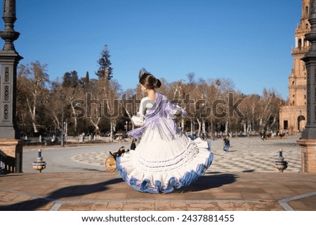 a little girl dancing flamenco dressed in a beige dress with ruffles and purple fringes in a famous square in seville, spain. The girl has flowers on her head and her hair in a bun