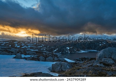 dramatic sunset over an idyllic mountain lake with ice on the water and snow covered peaks in the background along the Lysebotn road on a sunny day in Norway.