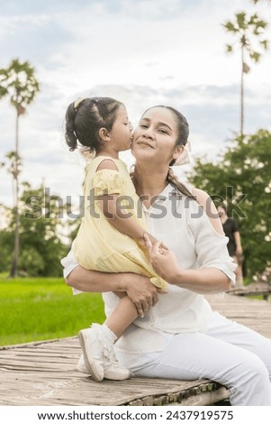 Asian mother and daughter express their love for each other by kissing her mother on the cheek on a wooden bridge in a rice field. Concept of giving love to family.