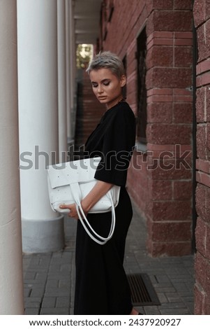 Side view of young female model in minimalist black dress carrying white leather bag and looking down outside, summer fashion street photo. 2024 trend influencer image, copy space. Red brick wall