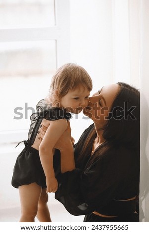 Happy mom hugging her child girl and kissing near window. The concept of cheerful childhood and family. Beautiful Mother and her baby. Copy space for text Royalty-Free Stock Photo #2437955665