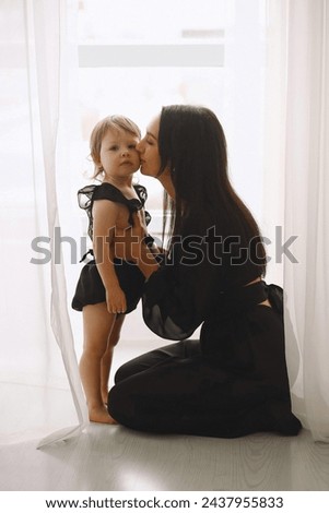 Happy mom hugging her child girl and kissing near window. The concept of cheerful childhood and family. Beautiful Mother and her baby in black outfits Royalty-Free Stock Photo #2437955833