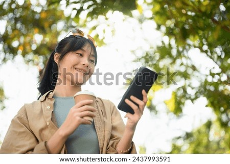 Satisfied Asian woman drinking coffee from paper cup using mobile phone in the park