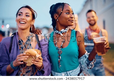 Happy female festival goers eating hamburger and drinking beer during open air music concert in summer. 