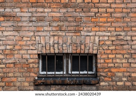 old red brick wall with a window with a white frame and metal bars. a bricked-up window in an old wall made of red brick of a different shade with a grille for ventilation