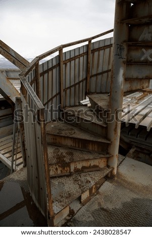 Weathered metal stairs spiral upward inside an industrial structure, hinting at the passage of workers past. The wear and rust speak to the harsh conditions and the relentless passage of time. Royalty-Free Stock Photo #2438028547