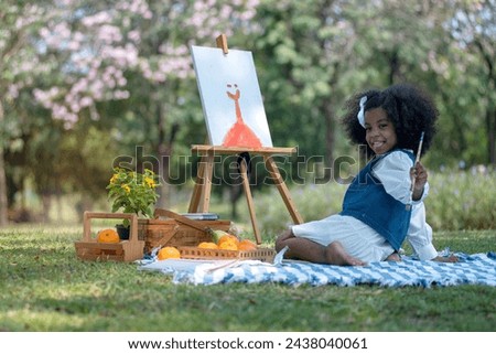 African little girl holds brush and paints picture on easel at park, very happy and smiling at her work, spend the holidays picnicking and painting