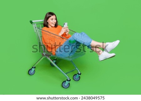 Full size portrait of impressed cheerful girl use smart phone inside hypermarket trolley isolated on green color background