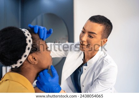 Facial aesthetics surgery consultations at medical beauty clinic. Young woman professional aesthetician examining patient's face, touching it with gloved hands. Royalty-Free Stock Photo #2438054065