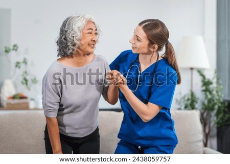 Caucasian female doctor assists in supporting an elderly Asian patient while they both sit on the sofa, ensuring comfort and stability during the interaction. Royalty-Free Stock Photo #2438059757