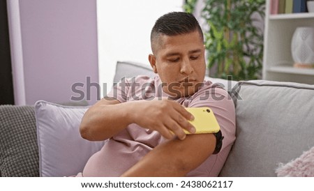 Confident latin guy, scanning his diabetes sensor with phone, monitoring glucose levels at home, serious yet relaxed Royalty-Free Stock Photo #2438062117