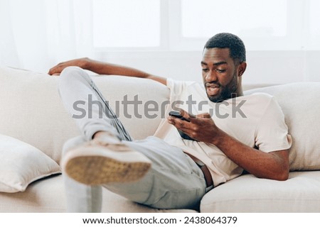Man sitting on a black sofa, happily typing messages on his smartphone He is enjoying the comfort of his modern apartment, using technology to stay connected and engaged This image captures the