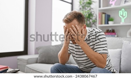 Stressed out young caucasian man sitting alone on a sofa at home, grappling with depression, frustrated and upset amid a personal crisis. Royalty-Free Stock Photo #2438067467
