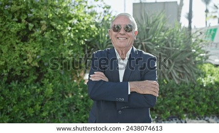 Cheerful senior man with grey hair, smiling joyously in the vibrant city park, elegantly sporting a suit and sunglasses, standing with a positive posture, arms confidently crossed. Royalty-Free Stock Photo #2438074613