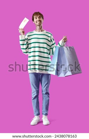 Young tattooed man with gift card and shopping bags on purple background