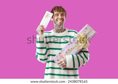 Young tattooed man with gift card and present on purple background