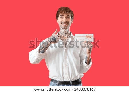 Young tattooed man pointing at gift card on red background
