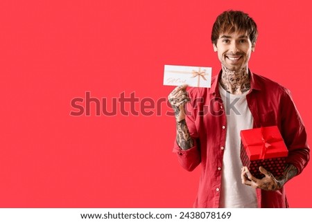 Young tattooed man with gift card and present on red background