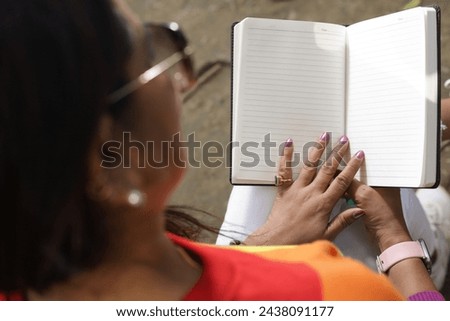 Portrait of mid aged woman holding a black color almanac in hand, taking notes in personal almanac. sunlight, green park environment. Royalty-Free Stock Photo #2438091177