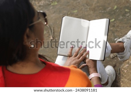 Portrait of mid aged woman holding a black color almanac in hand, taking notes in personal almanac. sunlight, green park environment. Royalty-Free Stock Photo #2438091181