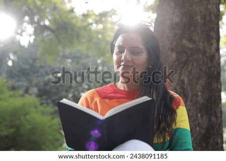 Portrait of mid aged woman holding a black color almanac in hand, taking notes in personal almanac. sunlight, green park environment. Royalty-Free Stock Photo #2438091185
