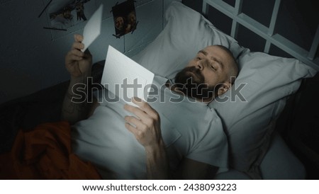 Male prisoner in orange uniform lies on bed in prison cell, looks at pictures with family and children. Criminal serves imprisonment term for crime in jail. Detention center or correctional facility.