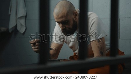 Inmate in tattoos in orange uniform sits on bed in jail cell, eats prison food from iron bowl. Male prisoner serves imprisonment term for crime in prison. Detention center or correctional facility. Royalty-Free Stock Photo #2438093227