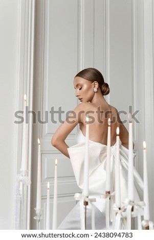 View from the back to a beautiful young woman in a white evening dress standing next to the candles in a white room. Royalty-Free Stock Photo #2438094783