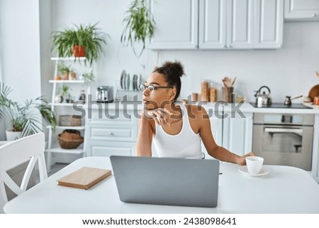 pensive african american woman in glasses sitting at desk with laptop and cup of coffee, remote work