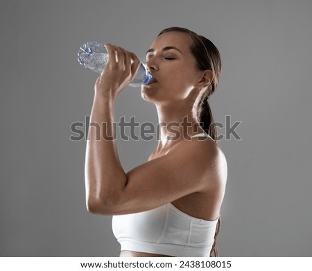 Woman, fitness and drinking with water for hydration after workout or exercise on a gray studio background. Thirsty female person or athlete with bottle of liquid for natural sustainability on mockup Royalty-Free Stock Photo #2438108015