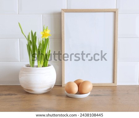 Spring still life. Blank picture frame mockup on wooden table background. Easter composition with daffodil flowers in a pot, hen eggs.  Farmhouse, Scandinavian interior.         