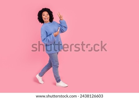 Full length photo of lovely young lady walk hold device show v-sign wear trendy blue knitted garment isolated on pink color background Royalty-Free Stock Photo #2438126603