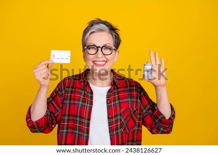 Photo of cheerful aged lady toothy smile hold debit card stack rolled dollar bills isolated on yellow color background