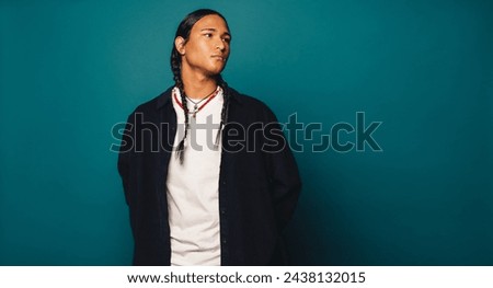 Confident Native American man with braided hair and ethnic necklace stands casually in a studio. His stylish appearance and self-assured demeanor display his cultural heritage and include him.