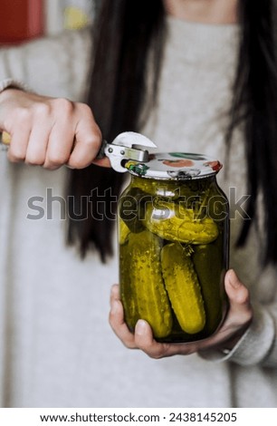 An adult woman, a gardener, a cook, holds in her hands in the kitchen a glass jar of vegetables with canned green cucumbers, preserves, opening the lid with an opener. Close-up photography of food. Royalty-Free Stock Photo #2438145205