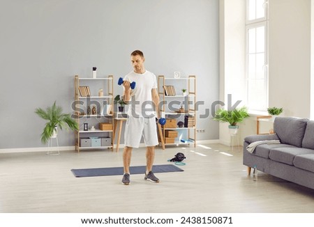 Ordinary man in morning in his home performs sports training and pumps his arm muscles. Young Caucasian man is standing in living room and using small dumbbells for sports workout. Home sports concept Royalty-Free Stock Photo #2438150871