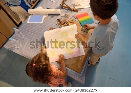 View from above of a teenage school boy and elementary age schoolgirl drawing on paper with pastel color pencils, creating a background for further during art class in the fine art school or gallery