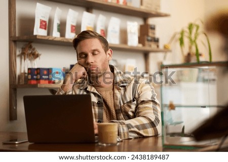 Portrait of bored freelancer, digital nomad sitting with laptop in cafe, leaning on his fist, looking with boredom at computer screen, drinking coffee, doing work task. Royalty-Free Stock Photo #2438189487