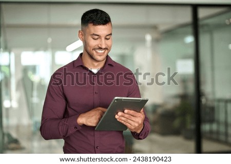 Happy hispanic businessman executive looking at tab device analyzing finance trading data working standing in office. Smiling busy young latin business man manager using tablet computer at work.