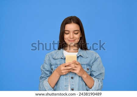Happy pretty gen z Latin teen girl, smiling teenage student with brunette hair wearing denim jacket holding smartphone, using cellphone looking at mobile phone standing isolated on blue background.