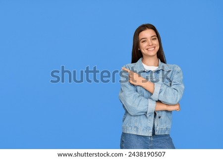 Happy cute Latin teenage student girl pointing aside standing on background. Smiling pretty hispanic teenager with brunette hair wearing denim jacket presenting ads standing isolated on blue.
