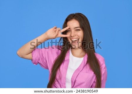 Happy pretty gen z Latin young woman, smiling freckled Hispanic brunette teenage girl wearing pink shirt having fun laughing showing peace sign over pretty face isolated on blue background. Portrait