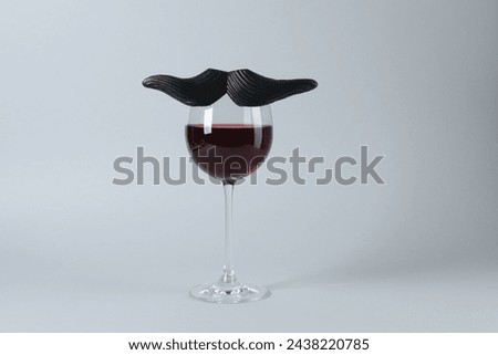 Glass of red wine with funny fake mustache on grey background